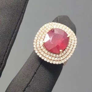 May include: A gold ring featuring a large, oval-cut ruby gemstone surrounded by two rows of small, sparkling diamonds. The ring is set on a gold band, creating a luxurious and eye-catching piece of jewelry.