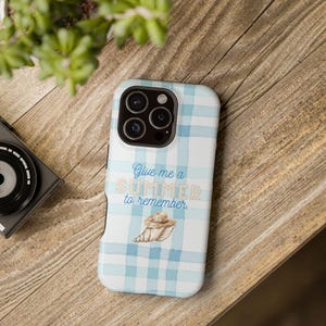 Puede incluir: Una funda de teléfono a cuadros blancos y azul claro con el texto "Give me a SUMMER to remember". La funda presenta un gráfico de un sombrero de paja y una concha. La funda del teléfono está sobre una superficie de madera.