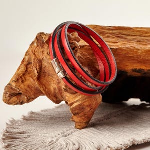 May include: A multi-strand bracelet with red and black leather bands. The bracelet has a silver-colored magnetic clasp. The bracelet is displayed on a piece of wood.