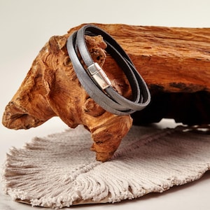 Puede incluir: Una pulsera envolvente de cuero gris con un cierre magnético plateado. La pulsera está envuelta alrededor de un trozo de madera texturizada de color marrón claro. El fondo incluye una tela beige con flecos.