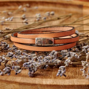 May include: A tan leather wrap bracelet with a silver-colored magnetic clasp. The bracelet is wrapped around itself and is resting on a wooden surface with dried lavender sprigs.