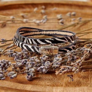 May include: A double-wrap bracelet featuring a black and white zebra stripe pattern on the exterior and a light tan interior. The bracelet is secured with a rectangular silver-toned metal magnetic clasp that connects the two ends of the strap.
