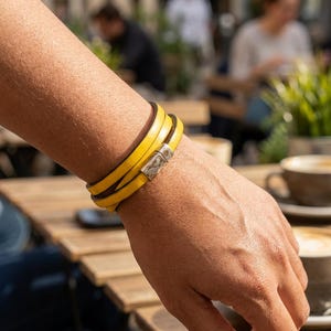 Op de afbeelding: Een felgele wikkelarmband met een gladde, leerachtige textuur en een rechthoekige, zilverkleurige magnetische sluiting. De armband wikkelt zich meerdere keren om de pols, wat zorgt voor een gelaagde uitstraling met een gepolijste metalen sluiting.