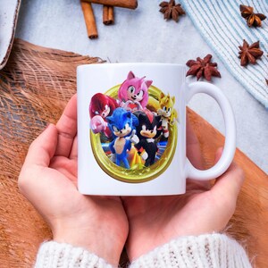 Pu&ograve; includere: Tazza in ceramica bianca con una grafica colorata di Sonic the Hedgehog e amici. Il design include Sonic, Tails, Knuckles, Amy Rose e Shadow, all'interno di una cornice dorata. La tazza ha un manico bianco.
