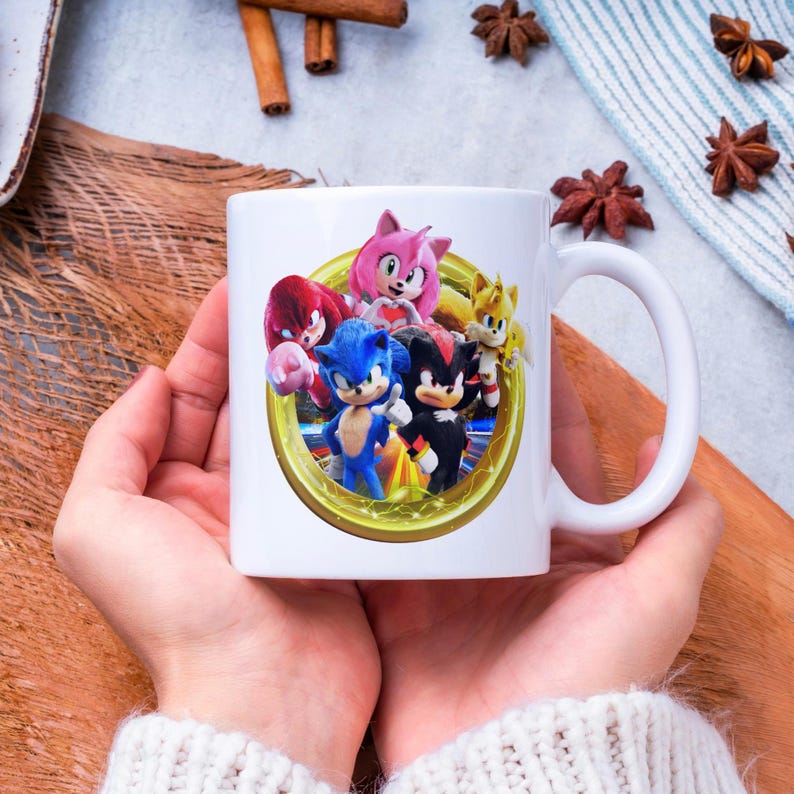 Pu&ograve; includere: Tazza in ceramica bianca con una grafica colorata di Sonic the Hedgehog e amici. Il design include Sonic, Tails, Knuckles, Amy Rose e Shadow, all'interno di una cornice dorata. La tazza ha un manico bianco.