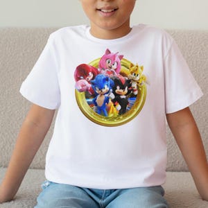 Pu&ograve; includere: T-shirt bianca a maniche corte con una grafica colorata di Sonic the Hedgehog e i suoi amici. Il design include Sonic, Tails, Knuckles, Amy Rose e Shadow, all'interno di una cornice circolare dorata.