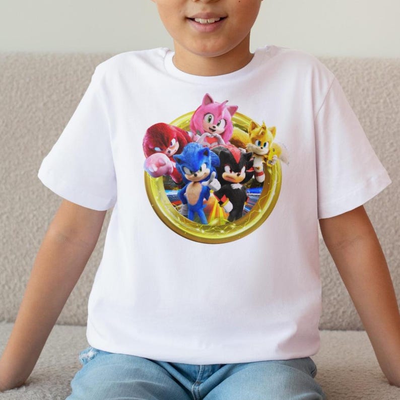 Pu&ograve; includere: T-shirt bianca a maniche corte con una grafica colorata di Sonic the Hedgehog e i suoi amici. Il design include Sonic, Tails, Knuckles, Amy Rose e Shadow, all'interno di una cornice circolare dorata.