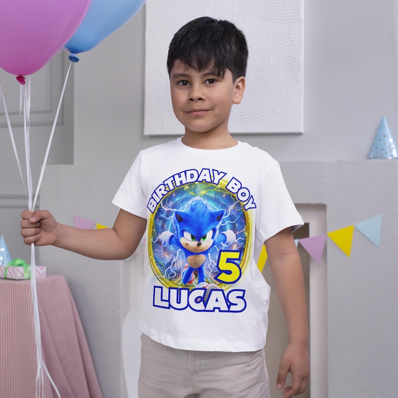 May include: White t-shirt featuring a blue Sonic the Hedgehog character graphic. The shirt reads "Birthday Boy" and "Lucas" with the number "5" in yellow.