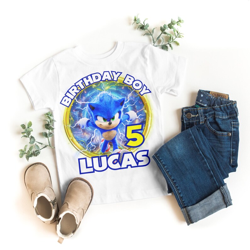 May include: White t-shirt featuring a blue Sonic the Hedgehog character graphic. The graphic includes a yellow ring with the text "Birthday Boy" and the number "5". The text "Lucas" is printed below the graphic.