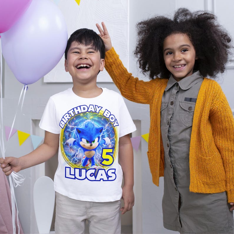 May include: A white t-shirt featuring a blue and yellow Sonic the Hedgehog graphic. The shirt reads "Birthday Boy" and "Lucas" with the number "5" in a yellow circle.