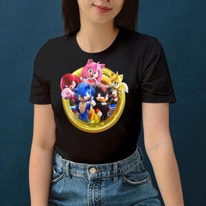 Pu&ograve; includere: T-shirt nera con una grafica colorata di Sonic the Hedgehog e amici all'interno di un anello d'oro. I personaggi includono Sonic, Tails, Knuckles, Amy Rose e Shadow. La maglietta &egrave; realizzata in materiale morbido.