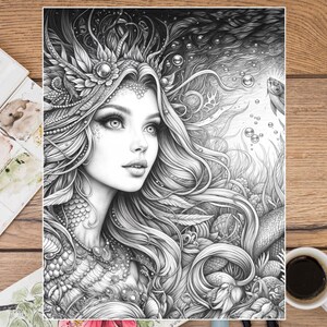 Puede incluir: Ilustraci&oacute;n en blanco y negro de una mujer con cabello largo y fluido, una corona detallada y joyas intrincadas. La obra de arte es una p&aacute;gina para colorear, con un tema de sirena, rodeada de plantas acu&aacute;ticas y burbujas. Una taza de caf&eacute; y una paleta de acuarela est&aacute;n en el fondo.