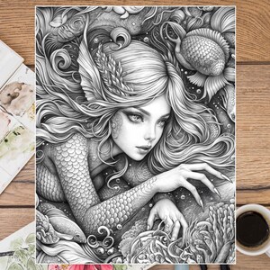 Puede incluir: Ilustraci&oacute;n en blanco y negro de una sirena con largo cabello suelto y escamas, rodeada de peces y plantas acu&aacute;ticas. La obra de arte detallada representa una escena submarina. Una taza de caf&eacute; y una paleta de acuarelas est&aacute;n en el fondo.
