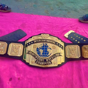 May include: A gold and black Intercontinental Heavyweight Wrestling Champion belt with a blue globe emblem. The belt has gold side plates with wrestling figures and is on a pink surface.