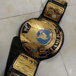 May include: A gold and black wrestling championship belt with a large, round center plate featuring a globe and the words "World Wrestling Federation Champion".