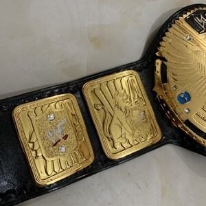 May include: A gold and black wrestling championship belt with a large central plate featuring a winged eagle design. The belt has three smaller plates, one with the WWF logo, one with a lion, and one with a globe.