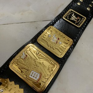 May include: A gold wrestling championship belt with black leather straps and a large gold plate featuring a lion design. The plate has a white and red shield with three stars on it. The belt also has a smaller gold plate with the letters "WWF" on it.
