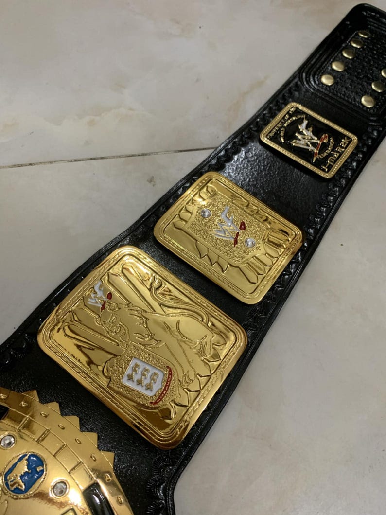 May include: A gold wrestling championship belt with black leather straps and a large gold plate featuring a lion design. The plate has a white and red shield with three stars on it. The belt also has a smaller gold plate with the letters "WWF" on it.