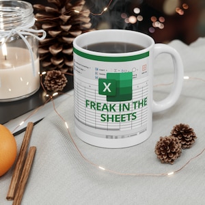 Può includere: Tazza in ceramica bianca con un design di foglio di calcolo Excel verde e bianco. La tazza ha un manico nero e il testo "Freak in the Sheets" è stampato sul davanti.