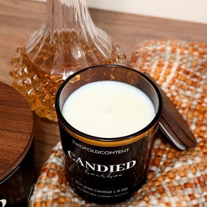May include: A black glass candle with the text "CANDIED bourbon" and a wooden lid. The candle is filled with white wax and sits on a textured orange and white fabric. An 8 oz soy wax candle.