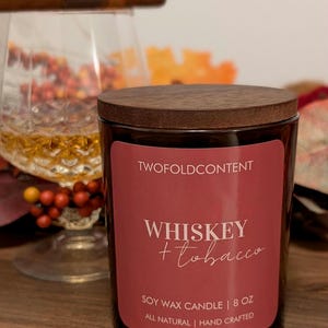 May include: A dark glass candle jar with a wooden lid. The label reads "Whiskey + Tobacco" and "Soy Wax Candle | 8 oz". A lit cigar and a glass of amber liquid are in the background.