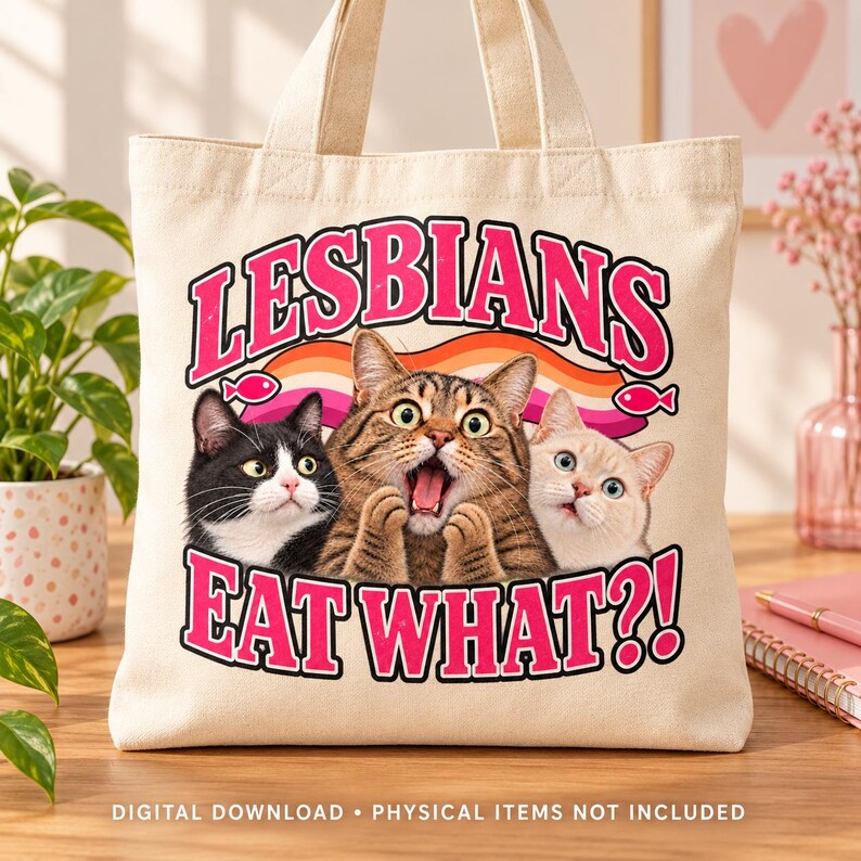 Cream canvas tote bag on a wooden table featuring three surprised cats, pink lettering, a lesbian pride ribbon, and two pink fish accents.