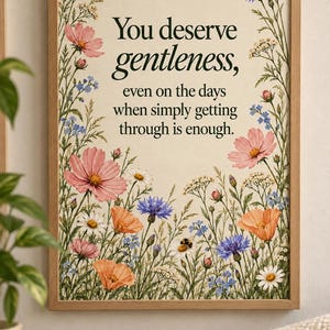 Wildflower affirmation print in a light wood frame on a cream wall beside a reading chair. Pink cosmos, white daisies, purple cornflowers, and blue forget-me-nots surround a centered quote about deserving gentleness.