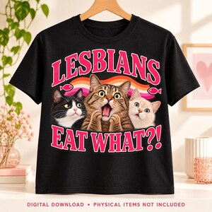 Black T-shirt on a wooden hanger featuring three expressive cats beneath pink “Lesbians” lettering, with “Eat What?!” below and a lesbian pride ribbon behind them.