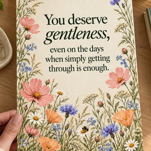 Unframed wildflower quote print held at one corner over a wooden desk. Forest-green lettering on ivory paper reads “You deserve gentleness, even on the days when simply getting through is enough,” surrounded by colorful flowers and a resting bee.