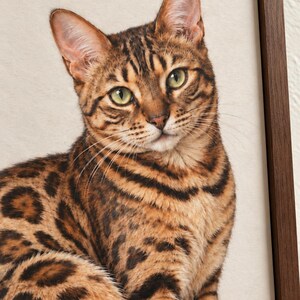 Close-up mockup of the printed Bengal cat’s green eyes, golden fur, dark facial markings, and fine whiskers, with a walnut frame edge visible on the right.