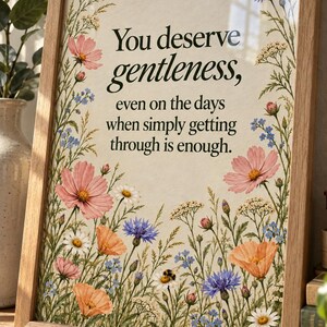 Angled view of a wildflower quote print in a wood frame on a shelf beside a plant. Natural light reflects softly on the glass, with forest-green lettering and colorful meadow flowers visible beneath.