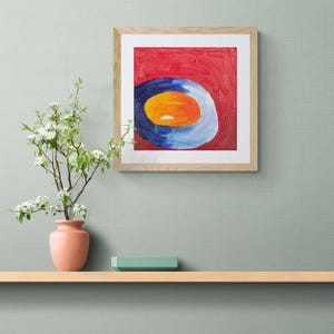 May include: A square framed painting featuring an abstract composition with a vibrant red background and a central circular motif layered with rings of deep blue, light blue, and a bright orange center, displayed with a peach-colored vase.