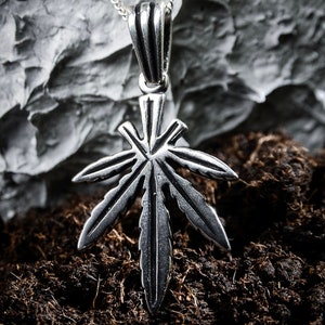 May include: A silver pendant in the shape of a marijuana leaf. The pendant is hanging from a silver chain and is resting on a dark brown surface.