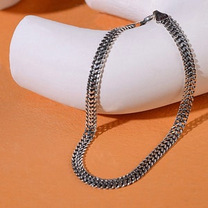 May include: A silver chain necklace with a woven design. The necklace is displayed on a white, curved surface against a warm, neutral background. The chain has a classic, timeless style.