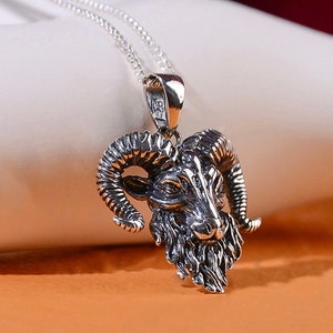 May include: Silver necklace with a detailed ram head pendant. The ram's head is facing forward with large horns and a textured beard.