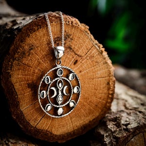 Op de afbeelding: Zilveren ketting met een ronde hanger met een ontwerp van maanfasen en een symbool van de drievoudige godin. De hanger ligt op een houten oppervlak.