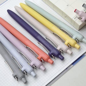 May include: A collection of Uni-ball ZENTO 0.5 pens in pastel colours, such as grey, lilac, coral, purple, and yellow. Each pen features a silver clip and the brand name printed on the barrel. The pens are arranged on squared paper.