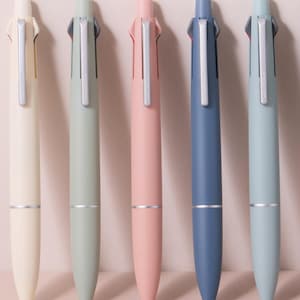 Könnte beinhalten: Fünf mehrfarbige Stifte in einer Reihe, jeder in einer anderen Pastellfarbe: Creme, Grün, Rosa, Blau und Hellblau. Jeder Stift hat einen silbernen Clip und einen silbernen Ring in der Nähe der Spitze. Die Stifte liegen auf einer hellrosa Oberfläche.