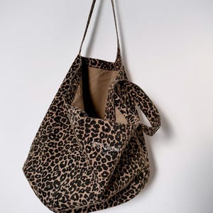 Könnte beinhalten: Eine Tragetasche mit Leopardenmuster und langen Trägern hängt an einem schwarzen, bärenförmigen Haken an einer weißen Wand. Die Tasche hat ein beiges Innenfutter und eine kleinere Tasche mit dem Text "LOCAL HONEY STUDIOS".