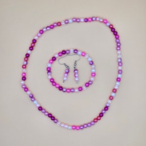May include: A jewellery set comprising a necklace, bracelet, and earrings. The set features small, round beads in shades of pink, purple, and white. The necklace and bracelet are circular, while the earrings dangle.
