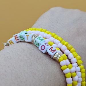 May include: A multi-layered bracelet with yellow and pink beads and white letter beads spelling out "BEST MOM".