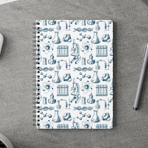 May include: A white spiral-bound notebook with a blue science-themed pattern, including beakers, microscopes, atoms, and DNA strands. The notebook is on a grey surface with a pen, earbuds, and other office supplies.