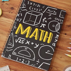 May include: A spiral-bound notebook with a black cover featuring white mathematical equations, geometric shapes, and the word MATH in large yellow letters. The notebook is surrounded by colored pencils, paper clips, scissors, and a ruler.