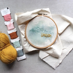 May include: A close-up of a white embroidery hoop with a watercolor-style London skyline design. The design includes the London Eye, Big Ben, and the words "London" in a gold thread. There are also several spools of embroidery floss in various colors.