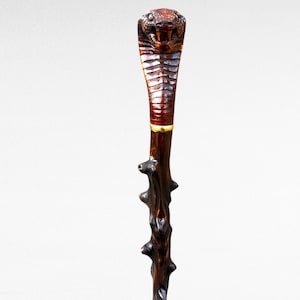 May include: A carved wooden walking stick featuring a cobra head. The head is detailed with scales and a fierce expression. The dark brown shaft has a textured, branch-like appearance, adding to its unique design.