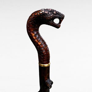 May include: A detailed walking cane with a snake head handle. The head is intricately carved, showing scales and a menacing open mouth. The cane's shaft is dark brown, with a gold-colored band. The overall design suggests a unique, handcrafted item.