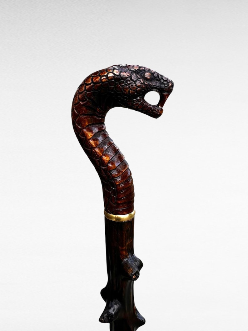 May include: A detailed walking cane with a snake head handle. The head is intricately carved, showing scales and a menacing open mouth. The cane's shaft is dark brown, with a gold-colored band. The overall design suggests a unique, handcrafted item.