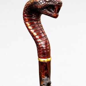 May include: A handcrafted wooden walking stick with a snake head handle. The handle is intricately carved with scales and a detailed open mouth. The shaft is dark brown with a gold band near the top. A unique accessory.