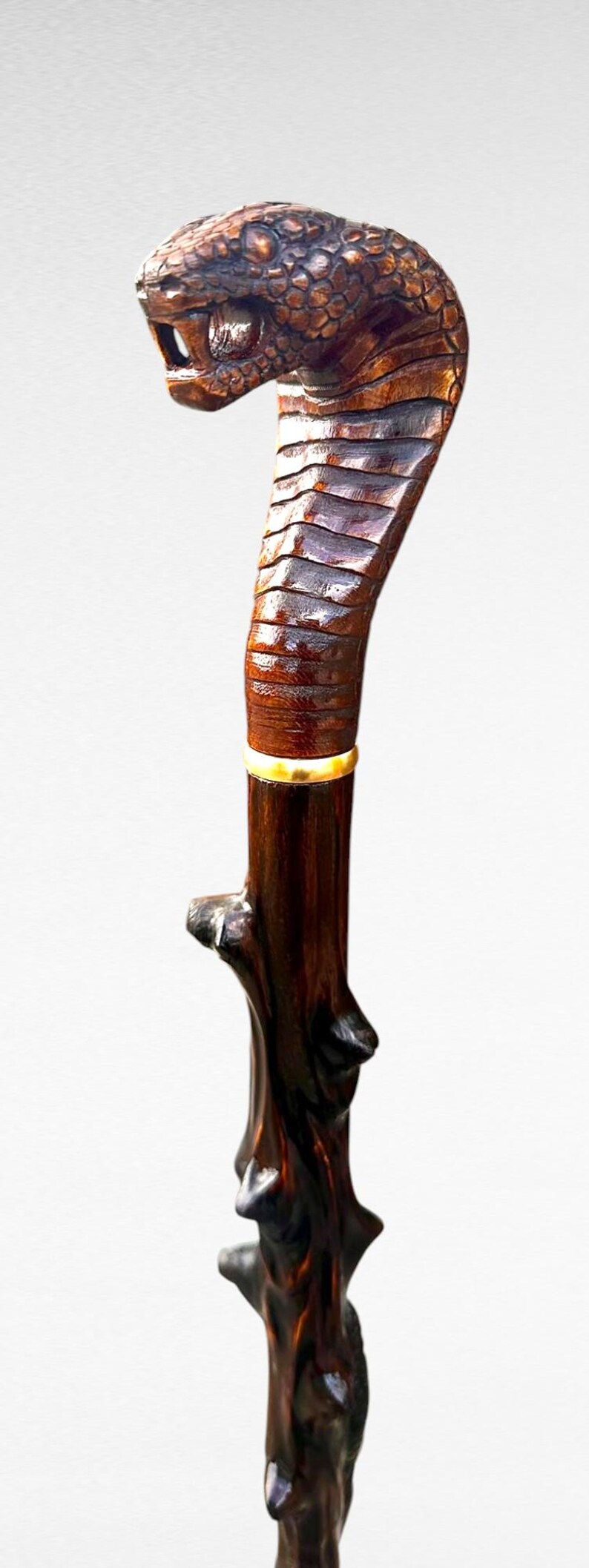 May include: A handcrafted walking cane featuring a cobra head handle. The handle is intricately carved from dark brown wood, with a detailed cobra head and a textured body. A gold-coloured band accents the shaft.