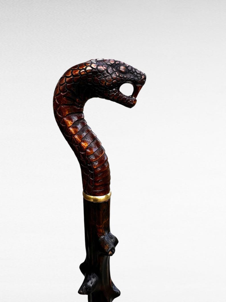 May include: A handcrafted wooden walking cane with a snake head handle. The snake head is intricately carved with detailed scales and open mouth. The cane is dark brown with a gold band near the top.